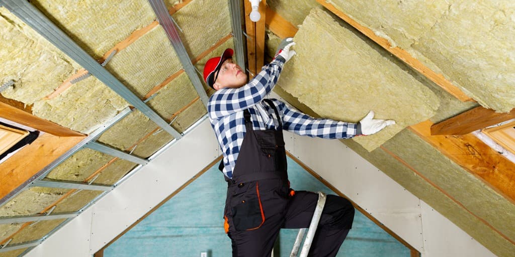 Technician installing yellow thermal insulation batts in a sloped ceiling with metal framing